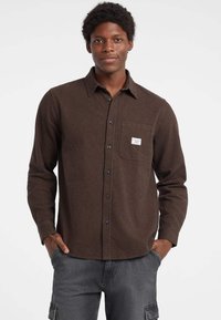 Camicia in flanella a scacchi marrone con maniche lunghe, tasca frontale, chiusura a bottoni e bottoni neri. Modello che indossa pantaloni grigi, mani in tasca.