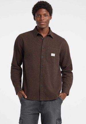 Camicia in flanella a scacchi marrone con maniche lunghe, tasca frontale, chiusura a bottoni e bottoni neri. Modello che indossa pantaloni grigi, mani in tasca.