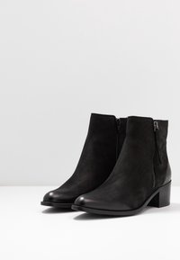 Botines de cuero negro que presentan una textura suave, punta redondeada, cremallera lateral y un tacón bajo de bloque.