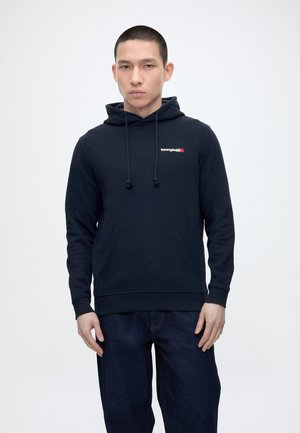 Młody mężczyzna w czarnej bluzie Tommy Jeans z przednią kieszenią i ciemnoniebieskich dżinsach, stojący na tle jednolitego jasnego tła.