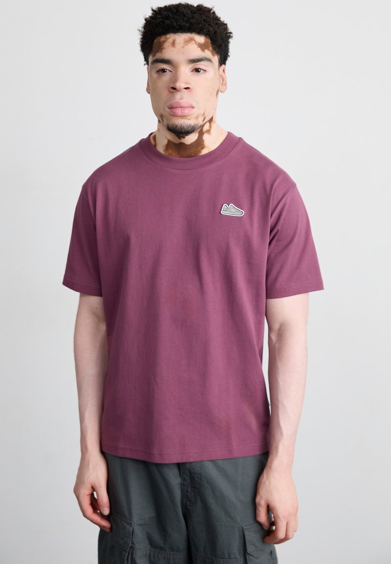 T-shirt in cotone bordeaux con maniche corte, presenta una piccola grafica di una scarpa sul lato sinistro del petto. Indossato con pantaloni cargo grigi.
