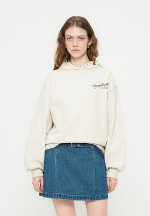 Νεαρή γυναίκα φοράει ένα ανοιχτό μπεζ φούτερ με το κείμενο «Sweet Goods» και μια μπλε τζιν φούστα με μαύρη ζώνη, στέκεται μπροστά σε λευκό φόντο.