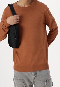 Osoba w brązowym swetrze i szarych spodniach cargo, trzymająca małą czarną torbę na ramieniu.