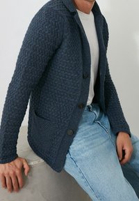 Marinblå texturerad cardigan med ett vävt mönster, enkel knappstängning och två fickor, i kombination med blå jeans på en sittande person.
