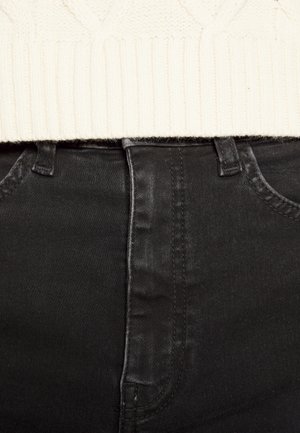 Gros plan sur la ceinture d'un jean en denim noir avec passants de ceinture et fermeture éclair frontale, associé à un ourlet de pull en maille crème.