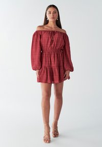 Rode off-shoulder jurk met lange mouwen, gestructureerde stof, getailleerde taille en geplooide rok. Gecombineerd met gouden sandaaltjes met bandjes.