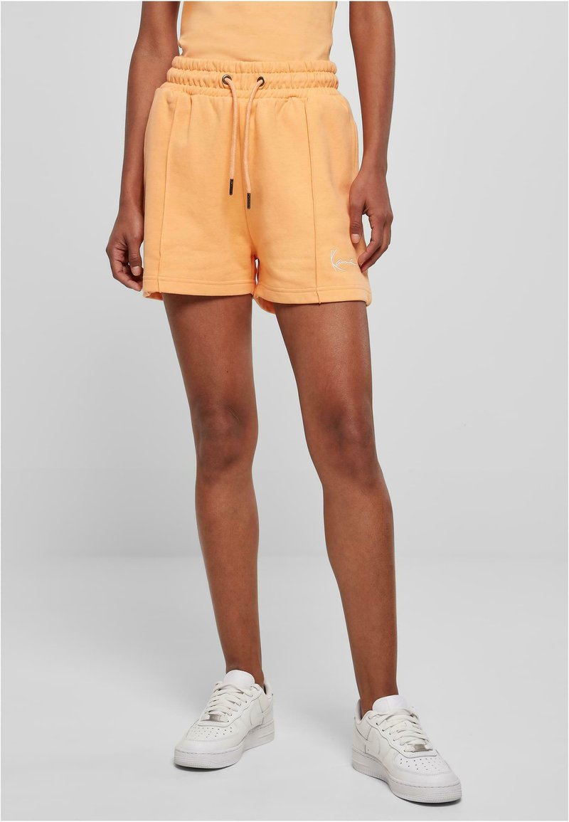 Lys oransje bomullsshorts med elastisk midje, snor, sidelommer og brodert logo på kanten. Kombinert med hvite joggesko.