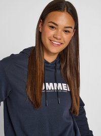 Smiling young woman with long straight hair wearing a dark blue hoodie with white text on the front against a plain background.