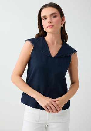Femme portant un haut bleu marine sans manches avec un large col et un pantalon blanc, debout les mains doucement jointes.