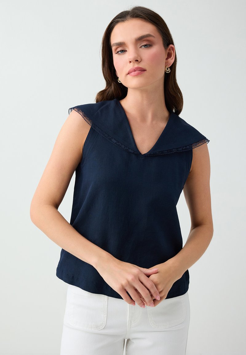 Femme portant un haut bleu marine sans manches avec un large col et un pantalon blanc, debout les mains doucement jointes.