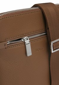 Borsa in pelle marrone con zip argentata, dettagli cuciti e tracolla in tessuto. Logo distintivo impresso sulla superficie in pelle.