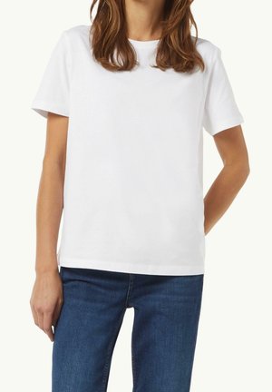 Kvinde iført en ensfarvet hvid kortærmet t-shirt og blå jeans, der står med den ene hånd afslappet og den anden bag ryggen.