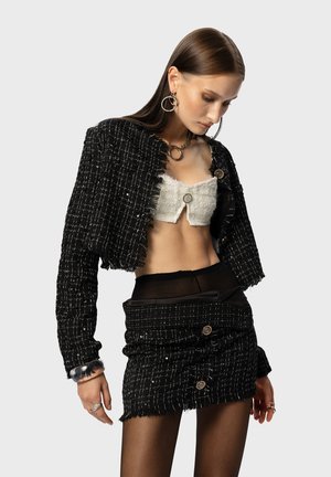 Black tweed cropped jacket with frayed edges, paired with a white textured top and black mini skirt featuring buttons. Accessories include large hoop earrings and a transparent bracelet.