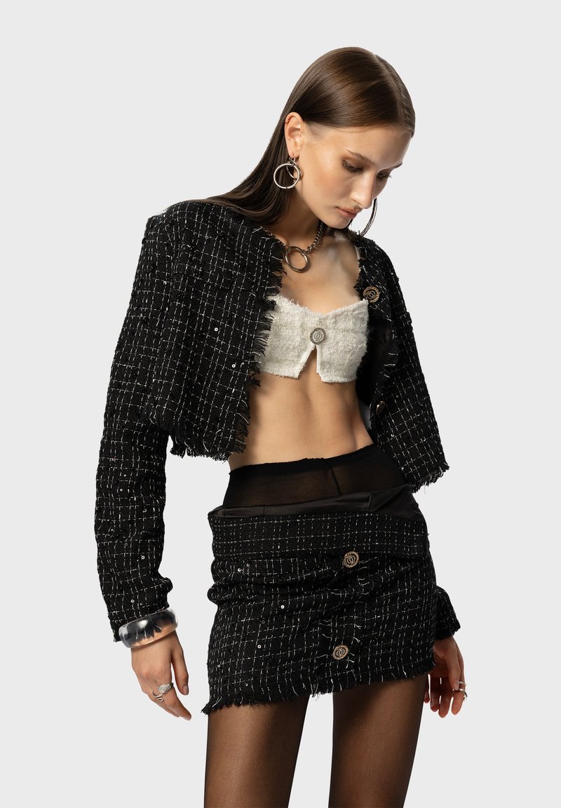 Giacca cropped in tweed nero con bordi sfrangiati, abbinata a un top bianco testurizzato e una mini gonna nera con bottoni. Gli accessori includono grandi orecchini ad anello e un braccialetto trasparente.