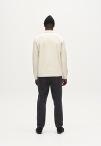 Világos bézs textúrájú pulóver hosszú ujjakkal, sötétszürke nadrággal és fekete cipővel párosítva. A modell háttal áll a kamerának.