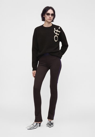 Femme portant des lunettes de soleil foncées, un pull noir avec le texte "Off-", un pantalon noir moulant avec une bande bleue et des talons pointus argentés, debout devant un fond blanc.