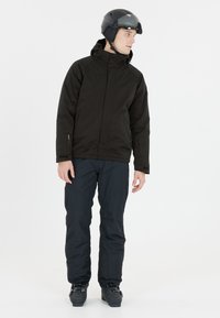 Schwarze isolierte Skijacke mit Kapuze, kombiniert mit dunkelblauen Schneehosen und schwarzen Skischuhen. Der Helm ist schwarz mit getönter Visier.