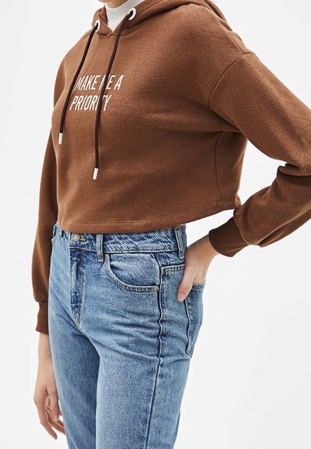 Persona con un abrigo corto marrón que dice "HAZME UNA PRIORIDAD" y jeans de tiro alto azules, con la mano en la cadera, contra un fondo blanco.
