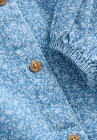 Blåt mønstret stof med hvide bladdesigns, naturlige træknapper og elastiske ærmekanter, der skaber teksturerede detaljer.