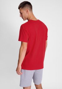 Rød bomulds T-shirt, korte ærmer, rund hals, båret med grå shorts. Stoffet ser blødt ud med en glat tekstur.