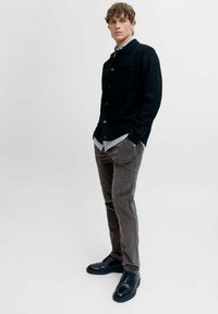 Cárdigan de punto negro con botones, combinado con una camisa de cuadros azul claro y blanca y pantalones de pana marrones. Unos zapatos negros completan el conjunto.