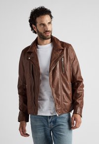 Brun lädermotorcykeljacka med en klämd krage, dragkedjefickor och en figursydd siluett, bärs över en vit t-shirt och jeans.