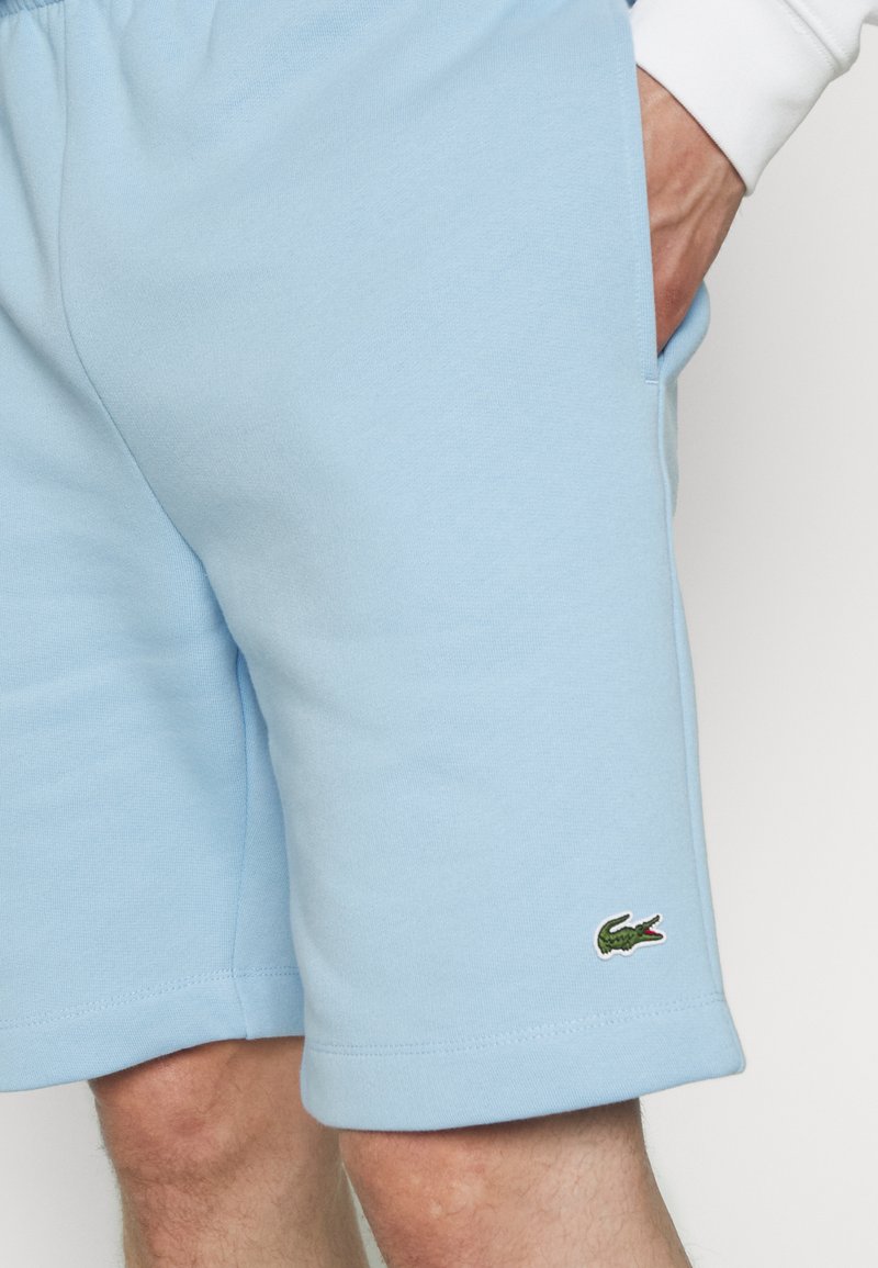 Shorts bleu clair en tissu doux, présentant une coupe décontractée et un logo de crocodile vert brodé sur l'ourlet gauche.
