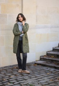 Lungo cappotto trapuntato verde oliva con sciarpa grigia, jeans scuri e stivali marrone chiaro; in piedi contro un muro di pietra chiara e un pavimento in ciottoli.
