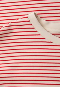 Gros plan sur un t-shirt crème à col rond avec de fines rayures horizontales rouges, uniformément espacées.