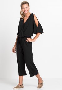 Combinaison noire à manches larges et découpes aux épaules, avec un devant croisé et un pantalon court. Assortie avec des chaussures à enfiler au motif léopard.