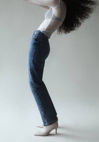 Personne portant des bottes blanches à talons, un jean bleu et un haut blanc à manches longues, se penchant en arrière avec les cheveux tombant vers le bas sur un fond uni.
