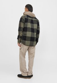 Camisa de franela a cuadros en negro y verde olivo, combinada con pantalones cargo beige y zapatos negros. Textura suave con un diseño con capucha.