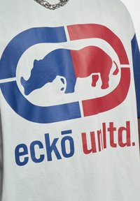 Balta trumpa rankovė marškinėliai su dideliu mėlynos ir raudonos spalvos rinokero grafinis logotipas ir tekstu "eckō unlt.d." priekyje. Matoma sidabrinė grandinėlė.