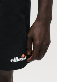 Σκούρο δέρμα χέρι ακουμπά κοντά στο τελείωμα μαύρων σορτς με το λευκό και πορτοκαλί λογότυπο ellesse σε απλό φόντο.