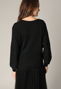 Pull en maille noir avec un décolleté large et une finition texturée, manches longues et coupe droite, associé à une jupe en tulle noir transparent.