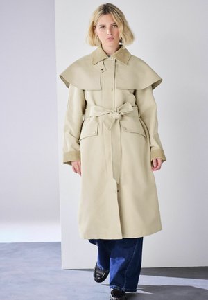 Blonde Frau, die einen beigefarbenen Trenchcoat mit Gürtel und weitem Kragen sowie blaue Jeans trägt, steht vor einem einfarbigen hellen Hintergrund.