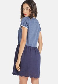 Mujer con cabello rizado oscuro que lleva una blusa azul y un vestido azul marino, mirando ligeramente hacia un lado, sonriendo y exhibiendo el dobladillo bordado del vestido.