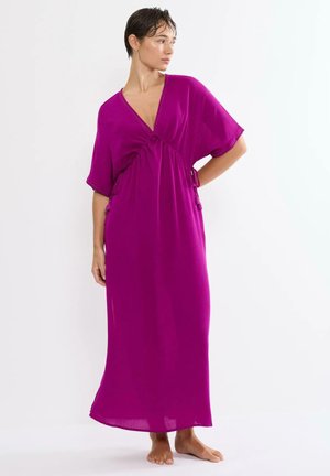 Vrouw staat blootsvoets in een losse, felpaarse, enkellange jurk met korte mouwen en een V-hals, kijkend naar links.