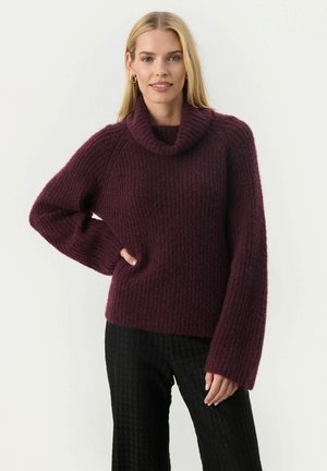 Burgundska pletena turtlenecka z visoko teksturo in sproščenim krojem. Ima široke rokave in prijeten, mehak videz.