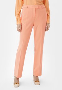 Spodnie w kolorze brzoskwiniowym, dopasowane, z prostymi nogawkami, gładką fakturą i plisami z przodu; uzupełnione dopasowaną pomarańczową koszulą.
