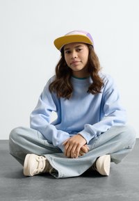 Himmelblaues Sweatshirt mit lockerem Schnitt, kombiniert mit hellblauen weiten Jeans und beigen Sneakers. Eine mehrfarbige Mütze rundet den Look ab.