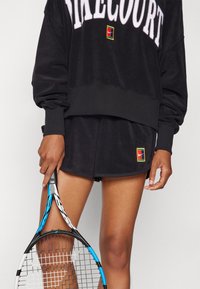 Must osaotsikuga must sweatshirt, millel on "FIRECOURT" tekst ja värvikas logo, sobib koos vastava musta lühikeste pükstega, millel on väike logo. Tennisereket käes.