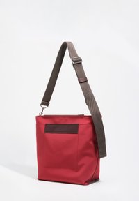 Czerwona torebka na ramię z regulowanym brązowym paskiem, przednią kieszenią i zamkiem błyskawicznym, prezentowana na jednolitym białym tle.