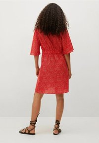 Femme aux cheveux bouclés portant une robe rouge à mi-mollet avec des pois blancs, posant de dos face à l'appareil photo, et des sandales plates en dentelle noire.
