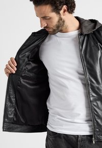 Svart läderjacka med mörk huva, dragkedjestängning och slät innervaddering; har en innerficka och avslappnad passform.