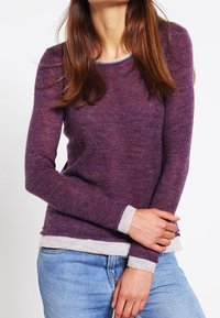 Fioletowy sweter dziany z okrągłym dekoltem, długimi rękawami i beżowymi lamówkami na mankietach oraz na dole. Wykonany z miękkiej, teksturowanej tkaniny. Noszony z niebieskimi dżinsami.