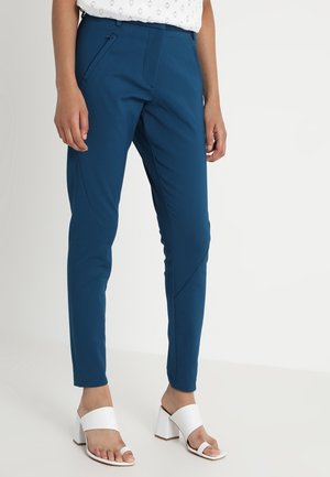 Frau trägt schlanke dunkelblaue Hose mit Reißverschlusstasche an der Seite und weiße Sandalen mit Blockabsatz, steht vor einem schlichten Hintergrund.