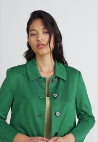 Green, fitted jacket with a collared neck, front button closure, and smooth texture. Buttons are dark with four holes, adding detail.