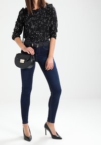 Blouse noire avec des taches blanches, manches volantes, associée à un jean skinny bleu foncé et des escarpins noirs à talon aiguille, portant un petit sac à main noir.
