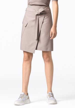 Jupe portefeuille beige avec poche latérale et ceinture nouée, portée avec des baskets blanches épaisses et un top côtelé sur une personne debout.